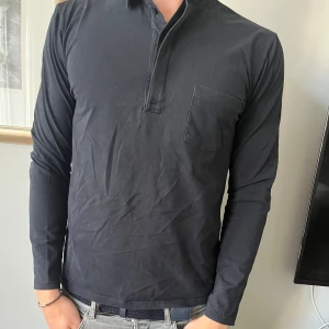 Svart långärmad pikétröja Flippa K - Svart långärmad pikétröja med klassisk krage och knappslå framtill. Tröjan har en bröstficka och är gjord i mjuk bomull, perfekt för en clean och stilren look. Passformen är normal och materialet känns skönt mot huden. Flippa K, skick 9/10, storlek M