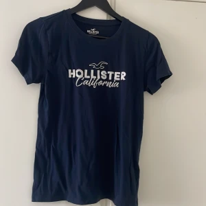 Mörkblå Hollister t-shirt med tryck - Snygg mörkblå t-shirt från Hollister med klassiskt vitt tryck 'Hollister California' och logga framtill. T-shirten har rund halsringning och korta ärmar. Perfekt basic-plagg med skön passform och mjukt bomullsmaterial.