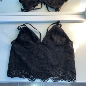 Svart spetsig linnetopp från BikBok - Svart linnetopp från BikBok med tunna, justerbara axelband och vacker spets över hela toppen. Blommigt spetsmönster och vågig spetskant nedtill ger en extra snygg touch. Perfekt att styla med höga jeans eller kjol för en trendig look. Har aldrig använt den 🥰
