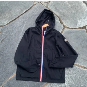 Svart vindjacka från Moncler uppe  för byten - Svart vindjacka från Moncler med huva och dragkedja i rött, vitt och blått. Jackan har Moncler-logga på ärmen, elastiska muddar och en clean, sportig look. Perfekt för dig som gillar stilrena och exklusiva plagg med streetkänsla.