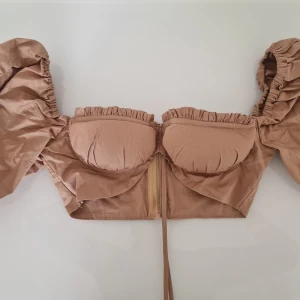 Beige korsettopp med puffärmar Lemon Lunar - Supersnygg beige korsettopp från Lemon Lunar med stora puffärmar och rynkade detaljer. Toppen har dragkedja bak, bygel vid bysten och knyt framtill. Perfekt för dig som vill sticka ut med en trendig och feminin look.