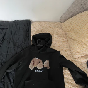 Svart Palm Angels hoodie med nalle - Svart hoodie från Palm Angels med ikoniskt nalletryck på bröstet och vit logga under. Klassisk huva och magficka framtill. Perfekt för dig som gillar streetwear och vill sticka ut med en unik detalj.