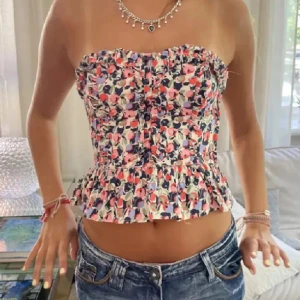 Blommig korsett topp - Säljer en blommig korsett topp med volangkant nertill och knappar framtill. Toppens färger går i rosa, blått, rött och vitt. Supersöt! Använd endast 2 gånger! Strl M men sitter som S