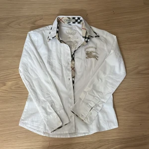 Vit skjorta med rutiga detaljer från Burberry - Säljer en klassisk vit skjorta från Burberry med ikoniska beige, svarta och röda rutiga detaljer på krage och ärmslut. Skjortan har broderad logga på bröstet och knäppning framtill. Perfekt för dig som gillar stilrena och tidlösa plagg. Äkta 