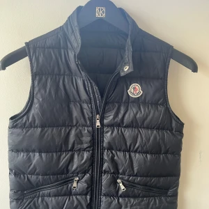 Svart dunväst från Moncler - Säljer en svart dunväst från Moncler med klassisk quiltad design och hög krage. Västen har dragkedja framtill, två fickor med dragkedja och Moncler-logga på bröstet. Perfekt för lager-på-lager under kyliga dagar.