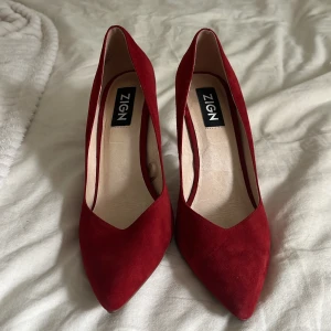 Röda klackar från Zign - Säljer ett par stilrena röda klackar från Zign med spetsig tå och hög klack. Skorna är i mocka och har en elegant form som passar perfekt till att lyfta vilken outfit som helst.