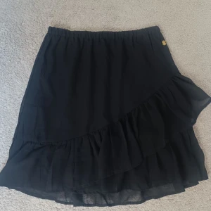 Svart volangkjol från Pernilla Wahlgren  - Säljer en svart kjol från Pernilla Wahlgrens kollektion på ullared med volangdetaljer och resår i midjan. Kjolen har en liten guldfärgad knapp med ananaslogga och är tillverkad i ett tunt, lätt material som ger ett flowy intryck. Det finns en underkjol som gör att den inte blir genomskinlig! Superfin och passar perfekt nu inför våren och sommaren. 100 kronor men priset kan diskuteras!! 