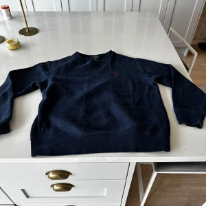 Mörkblå sweatshirt från Polo Ralph Lauren - Säljer en klassisk mörkblå sweatshirt från Polo Ralph Lauren med den ikoniska röda loggan broderad på bröstet. Tröjan har rund halsringning och långa ärmar. Perfekt för en stilren och avslappnad look. Storlek L i barn alltså 14-16y