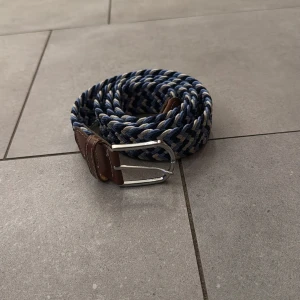 Blå Flätat bälte från onewear - Säljer ett snyggt flätat bälte i blått, vitt och beige med bruna skinn-detaljer vid spännet och änden. Bältet har ett silverfärgat metallspänne och passar perfekt till jeans eller chinos.
