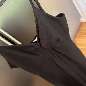 Svart långklänning med rosettdetalj - Säljer en svart långklänning med smala axelband och en snygg rosettdetalj framtill. Klänningen har en elegant och enkel design som passar perfekt till speciella tillfällen.