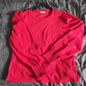 Röd långärmad topp från Cubus - Säljer en snygg röd långärmad topp från Cubus i storlek XL. Tröjan har puffiga axlar och smockade muddar vid ärmsluten. Materialet är lätt strukturerat.