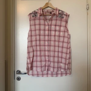 Ärmlös rutig skjorta med broderi - Säljer en ärmlös skjorta i bomull med rosa och svarta rutor samt broderade detaljer på axlarna och ryggen. Skjortan har klassisk krage och knäppning framtill. Perfekt för en avslappnad och cool stil. Från märket Odd Molly i Storlek 4 som enligt deras storleksguide motsvarar en storlek L/XL. Mycket fint skick, jag hittar inga synliga anmärkningar eller defekter. 