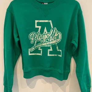 Grön sweatshirt med tryck från HM - Grön sweatshirt med stort vitt tryck på bröstet där det står 'Playoff' och ett stort A. Tröjan har rund halsringning, långa ärmar och ribbade muddar vid ärmslut och nederkant. Perfekt för en avslappnad stil.