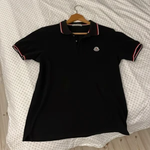 Svart pikétröja från Moncler - Säljer en svart pikétröja från Moncler med klassisk krage och knäppning framtill. Tröjan har röda och vita ränder på krage och ärmslut samt Moncler-logga på bröstet. Perfekt för en stilren och sportig look.