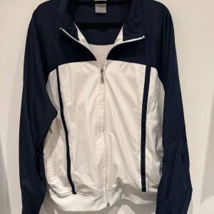 Vit och marinblå vindjacka med dragkedja - Säljer en sportig vindjacka i vitt och marinblått med hel dragkedja framtill och ståkrage. Jackan har långa ärmar och två fickor på sidorna. Perfekt för träning eller en aktiv vardag.