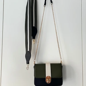 Grön och svart axelremsväska - Snygg axelväska i grönt och svart med vit detalj framtill och guldfärgat lås. Väskan har både en bred, justerbar axelrem och en tunn guldkedja. Perfekt för att lyfta din outfit med en modern touch. Köpt från Lindex. Priset kan diskuteras. 