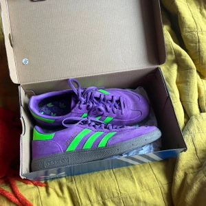  Adidas Handball Spezial W sneakers - Säljer ett par lila Adidas Handball Spezial W sneakers med gröna detaljer och snörning. Skorna har klassisk Adidas-design med tre ränder på sidan och platt sula. Perfekta för dig som vill sticka ut med färg på fötterna!