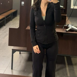Svart träningsjacka från Lululemon - Säljer en svart, figursydd träningsjacka från Lululemon med dragkedja framtill och två fickor med dragkedja. Jackan har hög krage och långa ärmar, perfekt för gymmet eller löprundan. Storlek 6.  (använt 2 gånger)