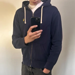 Mörkblå hoodie med dragkedja från Ralph Lauren - Mörkblå hoodie från Ralph Lauren med dragkedja framtill och vit dragsko i huvan. Klassisk gul logga broderad på bröstet. Mjuk bomullskänsla och två fickor framtill. Perfekt för en avslappnad stil. Det är i en tvättad mörkblå färg. Använd men inga skador. 