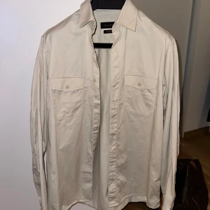 Beige overshirt från Massimo Dutti - Stilren beige overshirt, från Massimo Dutti i 100% bomull. Skjortan har två bröstfickor med knappar och klassisk krage. Perfekt för en clean och avslappnad look. Nypris 1 299 kronor. 