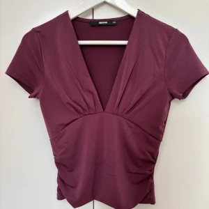 Vinröd v-ringad topp från Bik Bok - Säljer en vinröd topp från Bik Bok med djup v-ringning och korta ärmar. Toppen har rynkade detaljer framtill och är figurnära i modellen. Perfekt att styla med jeans eller kjol.
