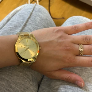 Guldig klocka från Michael Kors - Snygg och stilren klocka från Michael Kors i helguldigt utförande. Klockan har en rund urtavla med minimalistisk design och guldfärgade visare samt länkarmband i metall. Perfekt accessoar för att lyfta din outfit. Kostar 1400