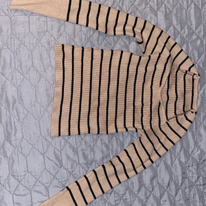 Beige och svart randig ribbad tröja - Säljer en beige ribbad tröja med svarta horisontella ränder. Tröjan har lång ärm och en något tight passform. Perfekt för dig som gillar klassiska mönster och enkel stil.