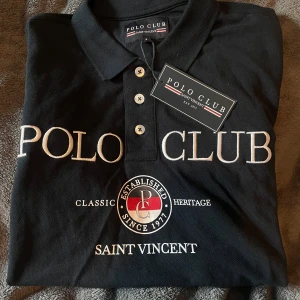 Svart pikétröja från Polo Club Saint Vincent - Klassisk svart pikétröja från Polo Club Saint Vincent med vit brodyr framtill och tre knappar vid kragen. Tröjan har korta ärmar och är perfekt för en stilren look. Märkeslogga och text på bröstet. Aldrig använt nypris 400kr