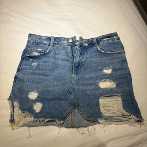 Blå jeanskjol - Säljer en blå jeanskjol från Zara med slitna detaljer och fransig nederkant. Kjolen har klassisk femficksdesign, knapp och dragkedja framtill.