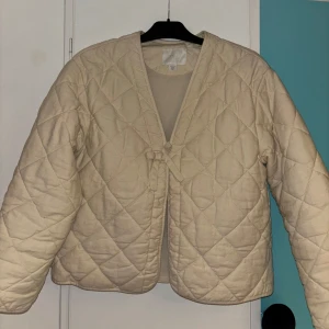 Krämvit quiltad jacka från Zara - Säljer en krämvit quiltad jacka från Zara med en söt rosett framtill. Jackan har lång ärm och en avslappnad passform. Perfekt för lager-på-lager och enkel att styla till olika outfits.🤍Skulle säga att storleken är xs/s