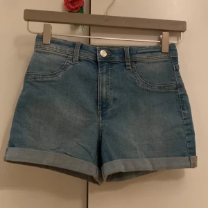 Blå jeansshorts med upprullad kant - Säljer ett par klassiska blå jeansshorts med upprullad kant och femficksmodell. De har normal passform och stängs med knapp och dragkedja. Perfekta för sommaren och enkla att matcha med olika toppar.