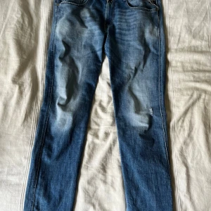 Blå Replay Anbass jeans slim fit - Säljer ett par blå Replay Anbass jeans i nästan nyskick med snygga slitningar framtill. Jeansen har dragkedja och knapp i midjan samt tydliga Replay-detaljer på bakfickorna. Slim fit, storlek W32/L32 