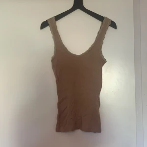 Beige ribbad linnetopp med spets från The Black Label Collection - Säljer en beige ribbad linnetopp från The Black Label Collection i storlek XS. Toppen har fina spetsdetaljer längs axelbanden och urringningen. Perfekt att styla till olika outfits!