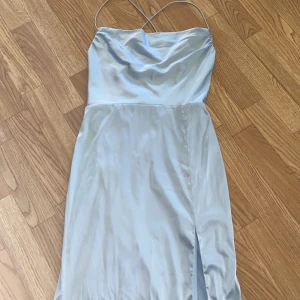 Ljusblå balklänning med korsade axelband - Säljer en ljusblå balklänning med tunna korsade axelband. Klänningen har en slits framtill och är tillverkad i ett glansigt satinliknande material. Från Nelly köptes för 800kr