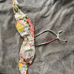 Blommig bikiniöverdel med knyt - Säljer en blommig bikiniöverdel med knyt i sidorna. Färgerna går i rosa, gul, grön och vit med ett romantiskt blommönster. Perfekt för sommardagar på stranden!