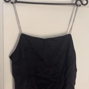 Svart festtopp med glittriga axelband - Säljer en svart topp med smala, glittriga axelband. Toppen har en enkel och stilren design med rak halsringning och figurnära passform. Aldrig använd 