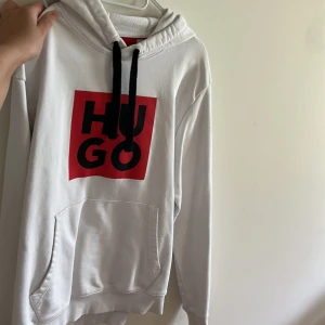 Vit hoodie från Hugo - Vit hoodie från Hugo med svart och röd logga framtill. Tröjan har huva med svarta snören och en stor magficka. Perfekt för en avslappnad stil.