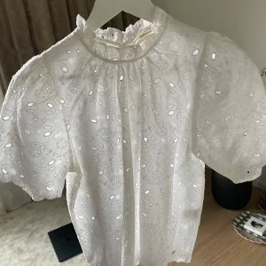Vit blus med broderade detaljer från Holly & Whyte - Superfin vit blus från Holly & Whyte med broderade blommor och hålmönster. Puffiga korta ärmar och liten volang vid halsen. Perfekt för en fräsch och somrig look!