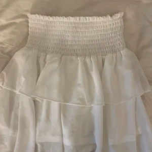 Vit volangkjol från Chelsea - Supersöt vit kjol från Chelsea med smock i midjan och flera lager volanger. Kjolen har en luftig och lätt känsla, perfekt för varma dagar. Passar dig som gillar romantisk och feminin stil. Använd fåtal gånger!!!