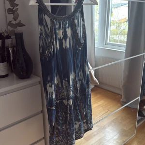 Blå mönstrad byxdress från Bobô Saint-Tropez - Säljer en blå och vit mönstrad klänning från Bobô Saint-Tropez. Shortsdress har ett bohemiskt paisleymönster, är ärmlös och har dekorativa detaljer vid halsringningen. Perfekt för sommardagar eller festivaler. Man kan spänna åt midjan!