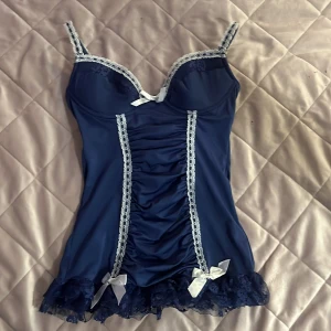 Mörkblå spetsprydd korsettopp med rosetter från Rampage - Säljer en mörkblå korsettopp från Rampage med vita spetsdetaljer och rosetter. Toppen har smala axelband, rynkade detaljer framtill och spetskant nertill. Perfekt för dig som vill ha något extra snyggt och unikt.