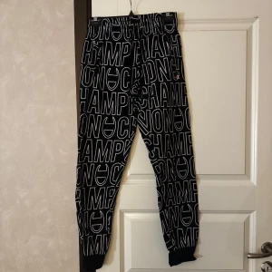 Svarta mjukisbyxor med vit text - Säljer ett par svarta mjukisbyxor med vit CHAMPION-text över hela byxorna. Byxorna har resår i midjan och vid bensluten, samt en avslappnad passform. Perfekta för chill eller träning.