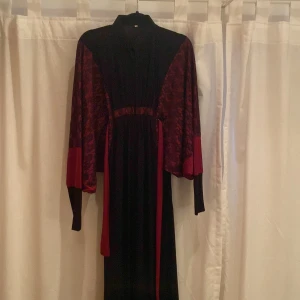 Svart och vinröd långklänning med spetsdetaljer - Säljer en elegant långklänning i svart med vinröda spetsdetaljer och vida ärmar. Klänningen har hög hals och markerad midja, samt kontrasterande vinröda partier på ärmarna. Perfekt för dig som vill sticka ut med en fin look.