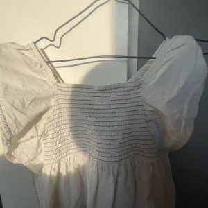Vit smockad tröja med puffärmar - Supersöt vit klänning med smockad överdel och puffiga korta ärmar. Perfekt för sommarens alla tillfällen och ger en romantisk känsla med sin luftiga design. Klänningen har en fyrkantig ringning och fina detaljer vid ärmslut.