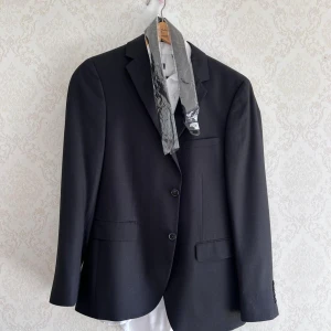 Kostymset från Dressmann med kavaj, skjorta och byxor - Säljer ett klassiskt svart kostymset från Dressmann i storlek M. Setet inkluderar en svart kavaj med två knappar, matchande svarta kostymbyxor och en vit skjorta och helt ny slips. Perfekt för formella tillfällen som bal eller nyår eller student. Allt i bilder ingår