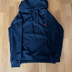 Svart hoodie från Ralph Lauren - Svart hoodie från Ralph Lauren med klassisk röd broderad logga på bröstet. Tröjan har huva med dragsko och en stor magficka framtill. Perfekt för en avslappnad och stilren look. Priset är diskuterbart