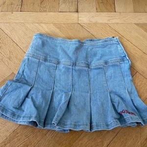 Ljusblå jeanskjol med veck - Säljer en ljusblå lågmidjad jeanskjol med veck och dragkedja i sidan. Kjolen har broderad röd text nertill och är i kort modell med en klassisk denimlook. Perfekt för dig som gillar en sportig och trendig stil. Den passar inte mig och har inte lyckats sälja pga oseriösa köpare💕