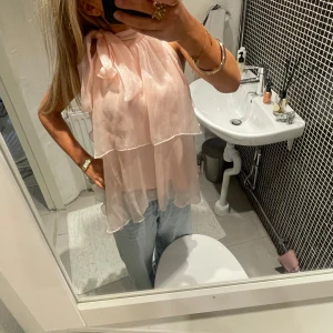 Ljusrosa volangblus med rosett - Säljer en ljusrosa blus med volanger och stor rosett vid halsen. Blusen är ärmlös och har ett luftigt, flowy material som ger en romantisk känsla. 