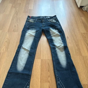 Mörkblå jeans med broderade detaljer - Säljer ett par mörkblå jeans från Young Age med slitningar framtill och broderade mönster på bakfickorna. Jeansen har normal passform och klassisk femficksdesign. Snygga kontrastsömmar och unika detaljer på fickorna.