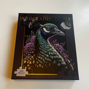 Holographic Puzzle med påfågelmotiv - Säljer ett holografiskt pussel med ett färgstarkt motiv av en påfågel omgiven av lila blommor. Pusslet består av 1000 bitar och kommer i en snygg svart kartong med gulddetaljer. Perfekt för dig som gillar utmanande pussel och vackra motiv. Pusslet är oöppnad, därav priset. 